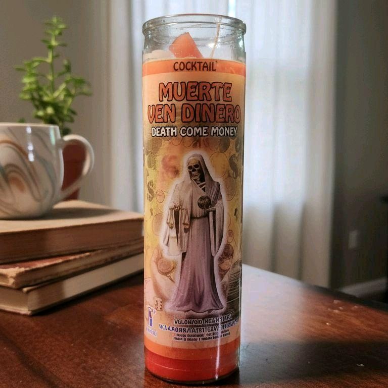 Veladora Muerte Ven Dinero naranja de siete días sobre mesa de madera con libros y taza